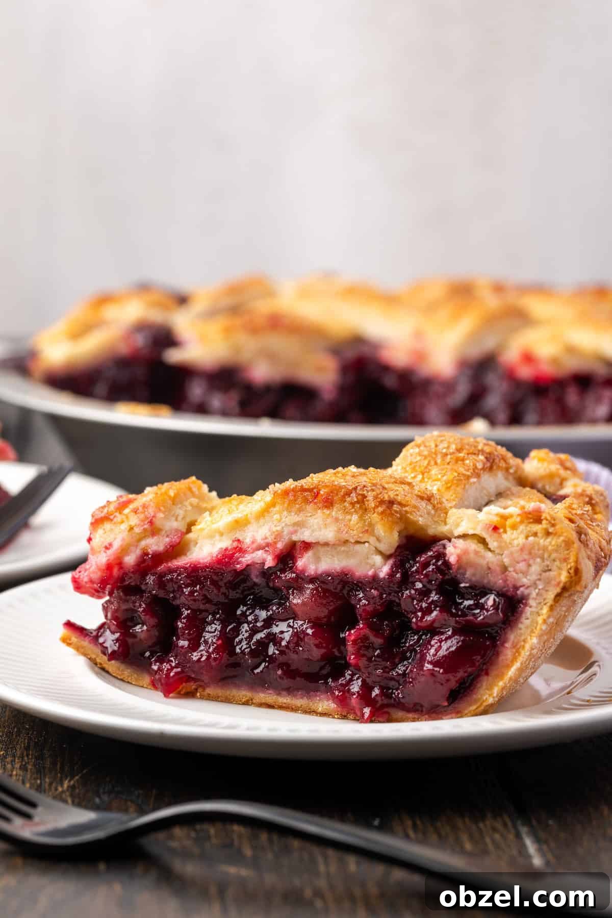 Easy homemade cherry pie with thick cherry filling and flaky pastry crust. Side view of a slice of cherry pie on a white plate with the rest of the pie in the background.