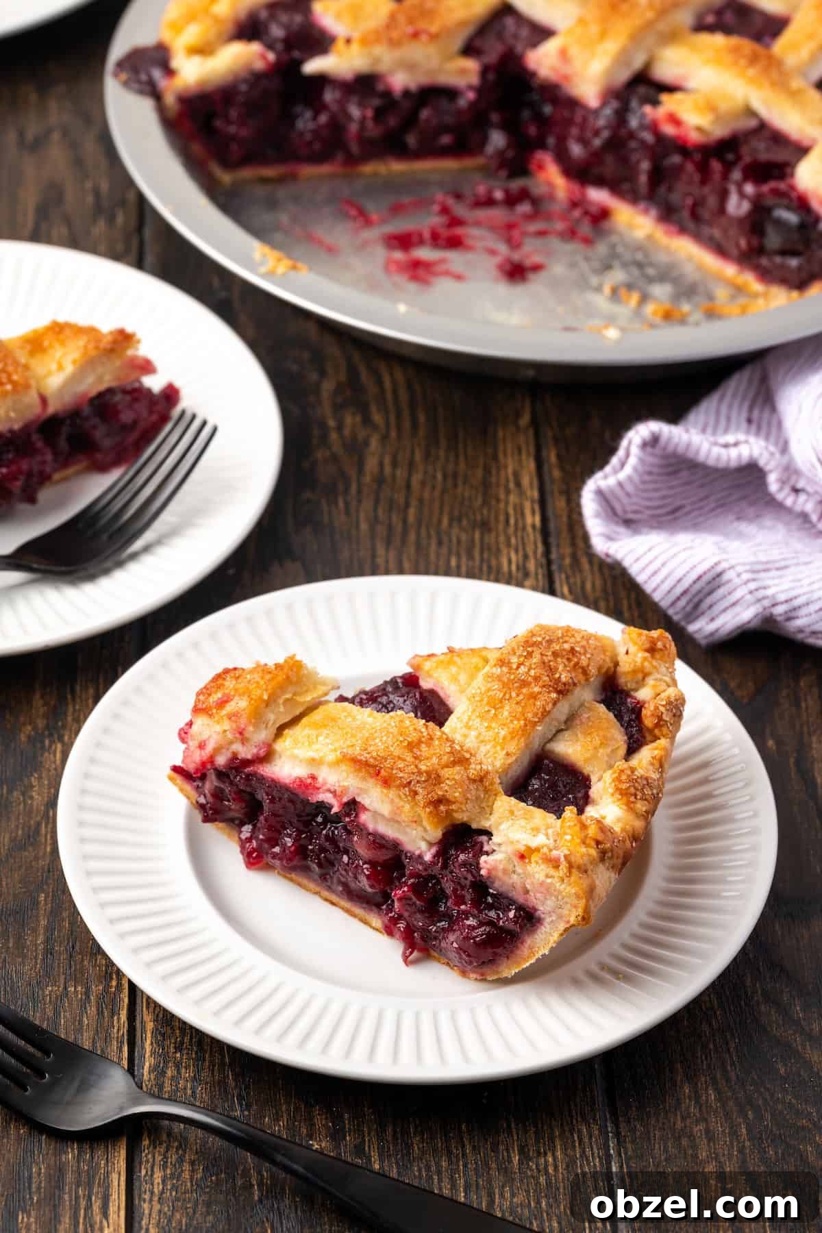 Slice of homemade cherry pie on a white plate. A slice of cherry pie on a white plate with the rest of the pie in the background.