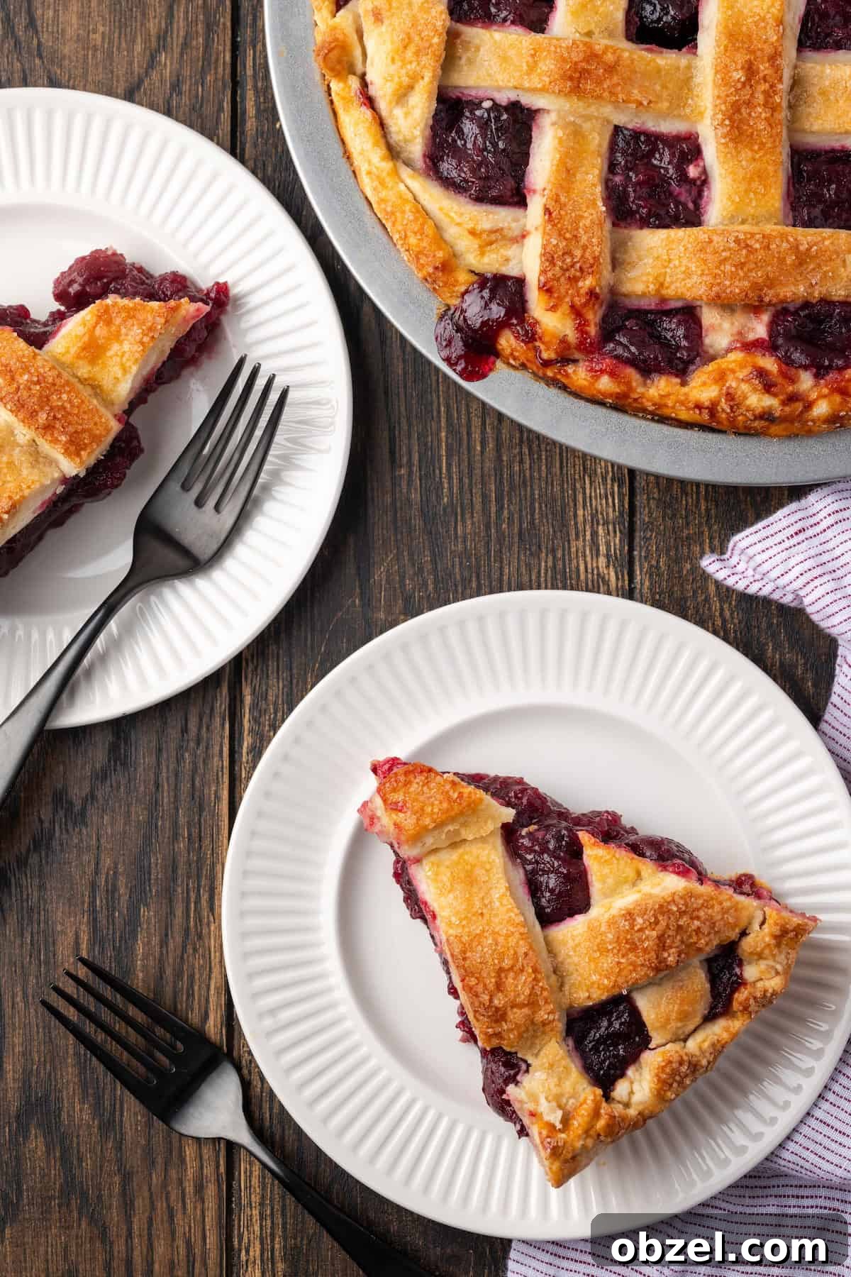 Thick slice of cherry pie with flaky crust. Overhead view of a slice of cherry pie on a white plate next to a second slice and the rest of the pie.