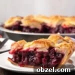 Side view of a slice of cherry pie on a white plate with the rest of the pie in the background.