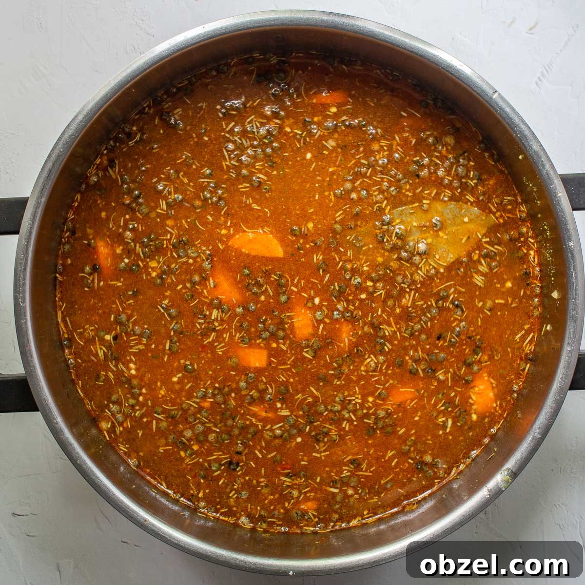 Traditional Spanish Lentil Stew with Chorizo 10 Uncooked dark lentils floating in a brown liquid with carrots and a bay leaf in a large steel saucepan.