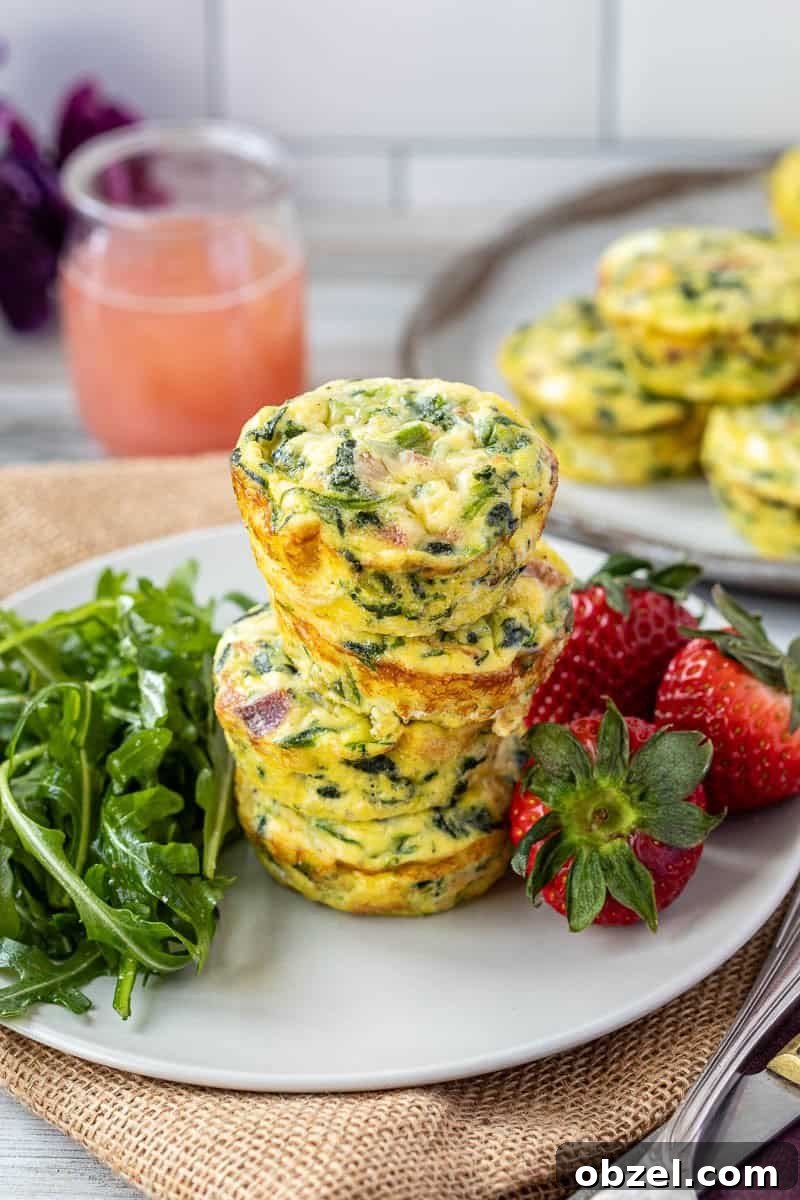 Mini Spinach Egg Bites: Oven-Baked Breakfast Recipe 2 Three spinach egg bites with bacon stacked on a plate with fresh arugula and strawberries, glass of pink juice in the background.