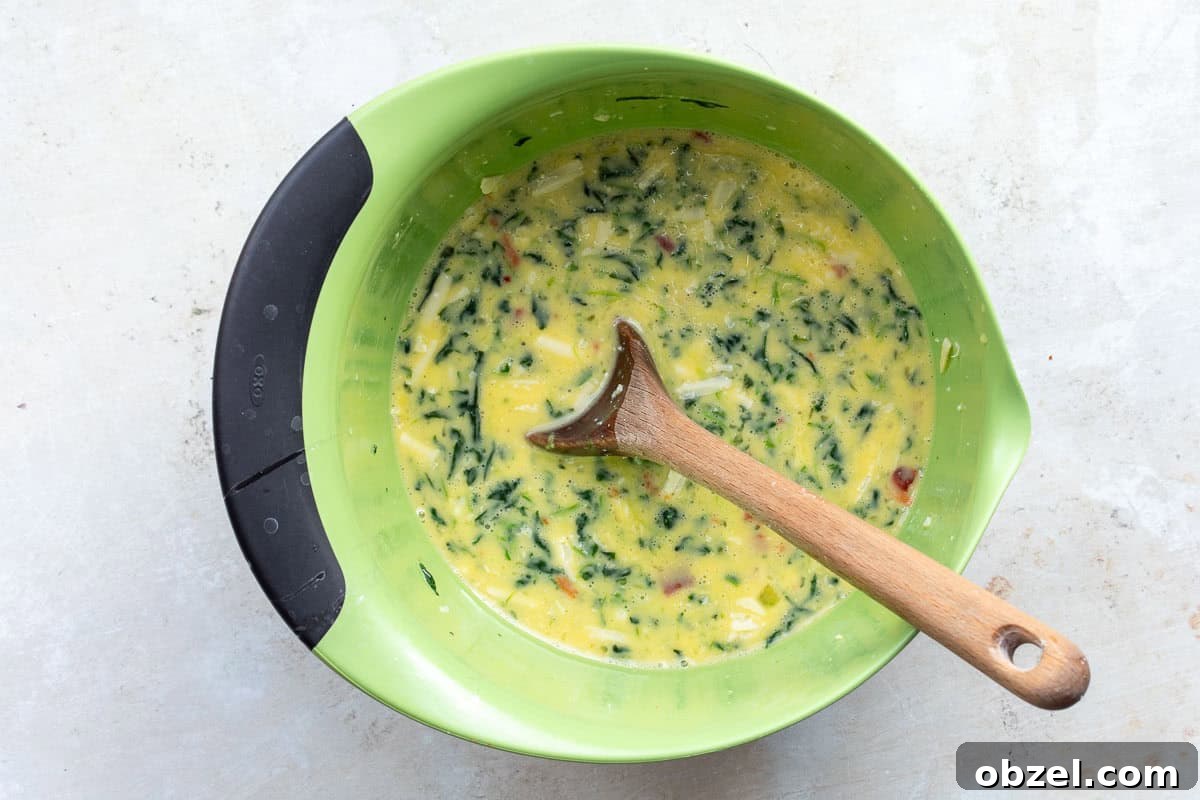 Mini Spinach Egg Bites: Oven-Baked Breakfast Recipe 6 A green mixing bowl contains a spinach, bacon, and egg mixture with visible herbs and spices. A wooden spoon rests inside the bowl.