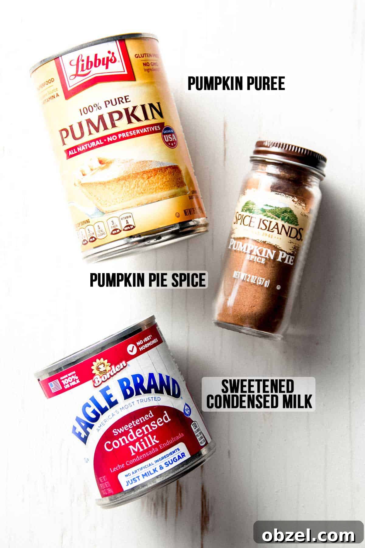 A can of pumpkin puree, sweetened condensed milk, and a jar of pumpkin spice sauce on a white wooden board.