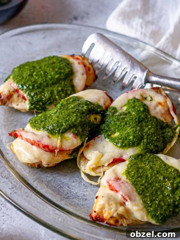 Close-up of air fryer Caprese chicken on a glass platter, showing melted cheese, juicy tomatoes, and vibrant basil pesto with a metal serving tong.