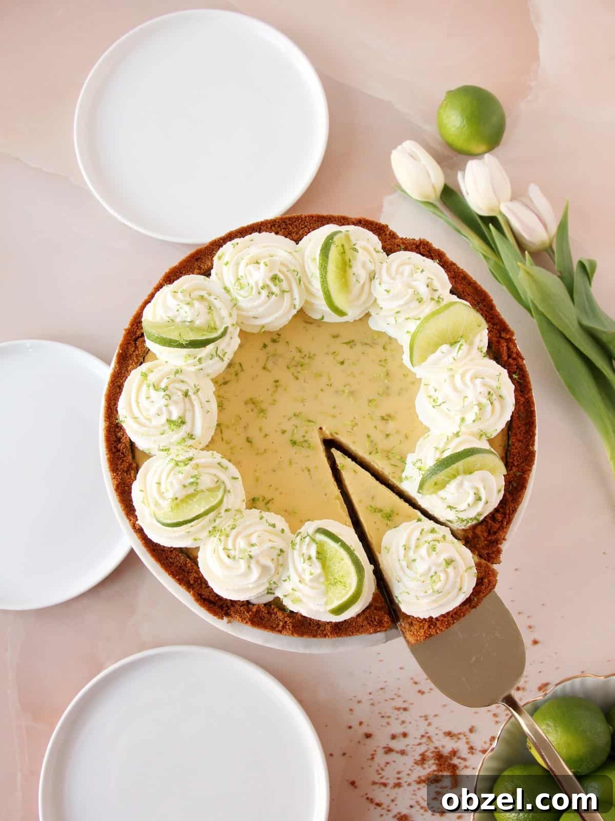 Key Lime Pie Recipe with Graham Cracker Crust 8 Keylime pie on cake stand, decorated with swirls of whipped cream, wedges of lime and lime zest. Three white round plates ready to be served. White tulips and limes as decoration on pink marble background.