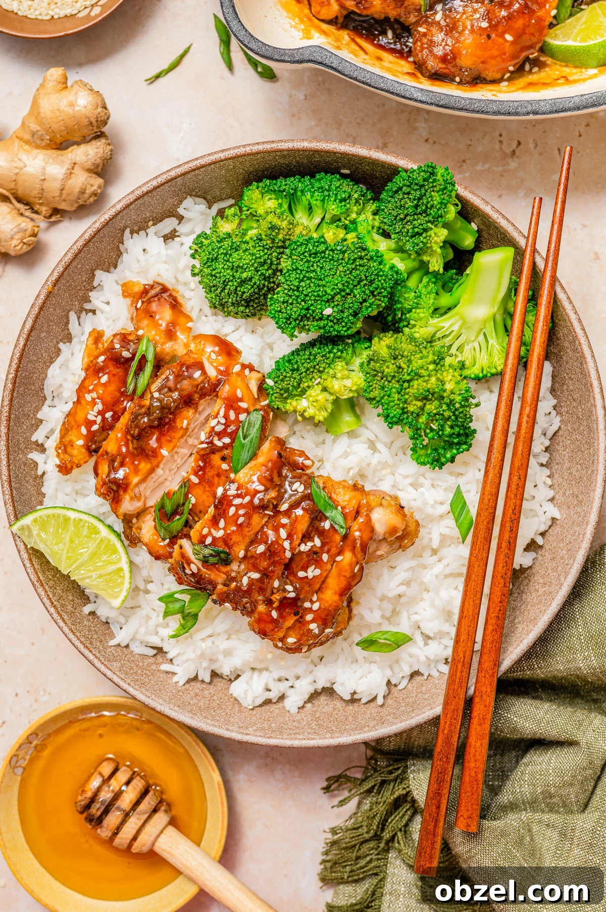 Sticky Honey-Ginger Chicken with Soy Glaze 2 A bowl of white rice topped with glazed chicken, broccoli florets, sliced green onions, sesame seeds, and a lime wedge, with chopsticks resting on the side.