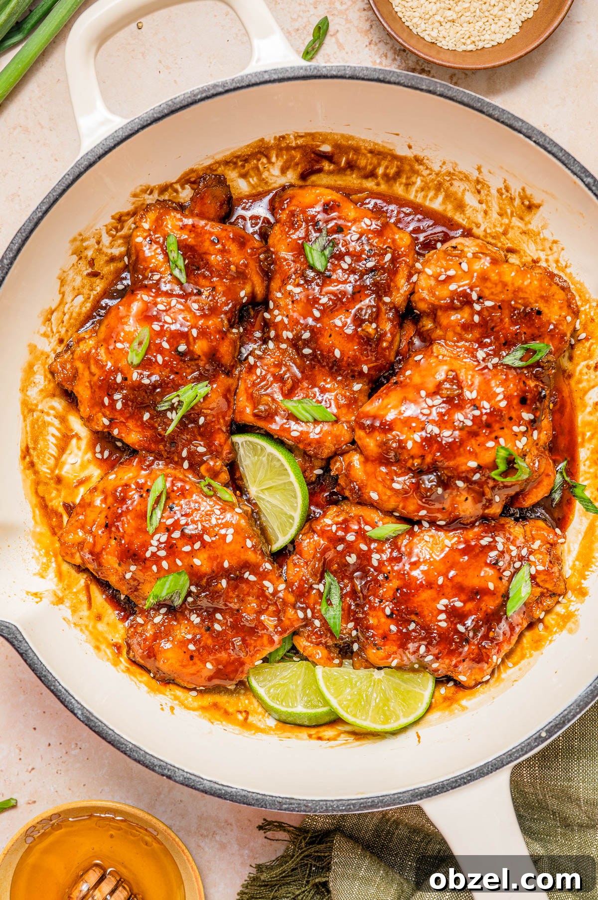 Sticky Honey-Ginger Chicken with Soy Glaze 3 A skillet filled with glazed chicken thighs topped with sesame seeds and sliced green onions, garnished with lime wedges.