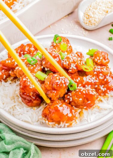 Plate of sesame chicken over rice with chopsticks, garnished with sesame seeds and green onions on a light tabletop.