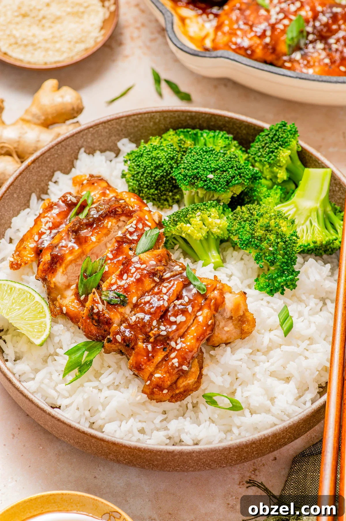 Sticky Honey-Ginger Chicken with Soy Glaze 5 A bowl of white rice topped with glazed chicken, sesame seeds, chopped green onions, steamed broccoli, and a wedge of lime.