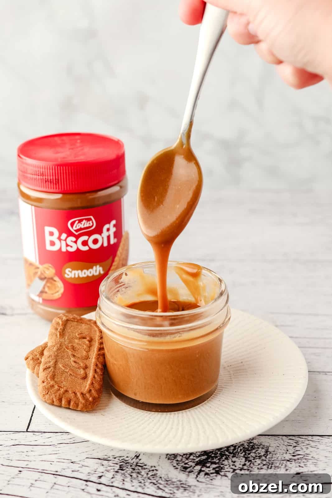 Biscoff Cookie Butter Sauce Recipe 2 A small mason jar of Biscoff sauce with a spoon full of sauce being drizzled into it, with Biscoff cookies on the side and jar of Biscoff spread in the background.