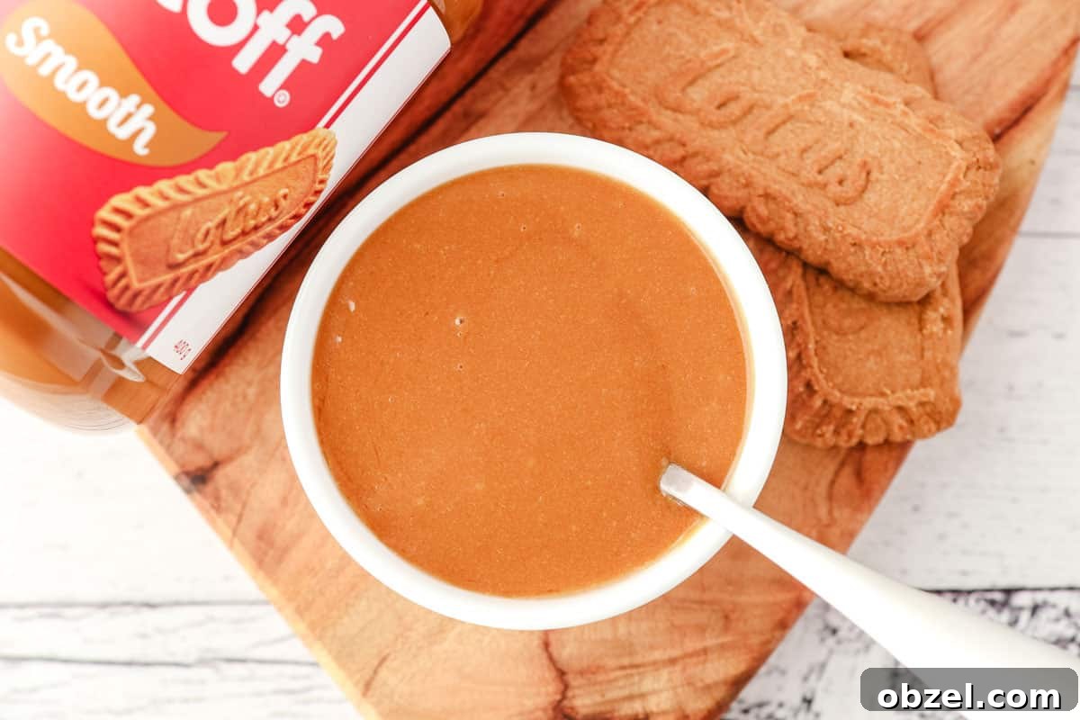 Biscoff Cookie Butter Sauce Recipe 7 Top down view of Biscoff topping in a serving bowl with a spoon in it, with Biscoff cookies and a jar of Biscoff spread on the side.