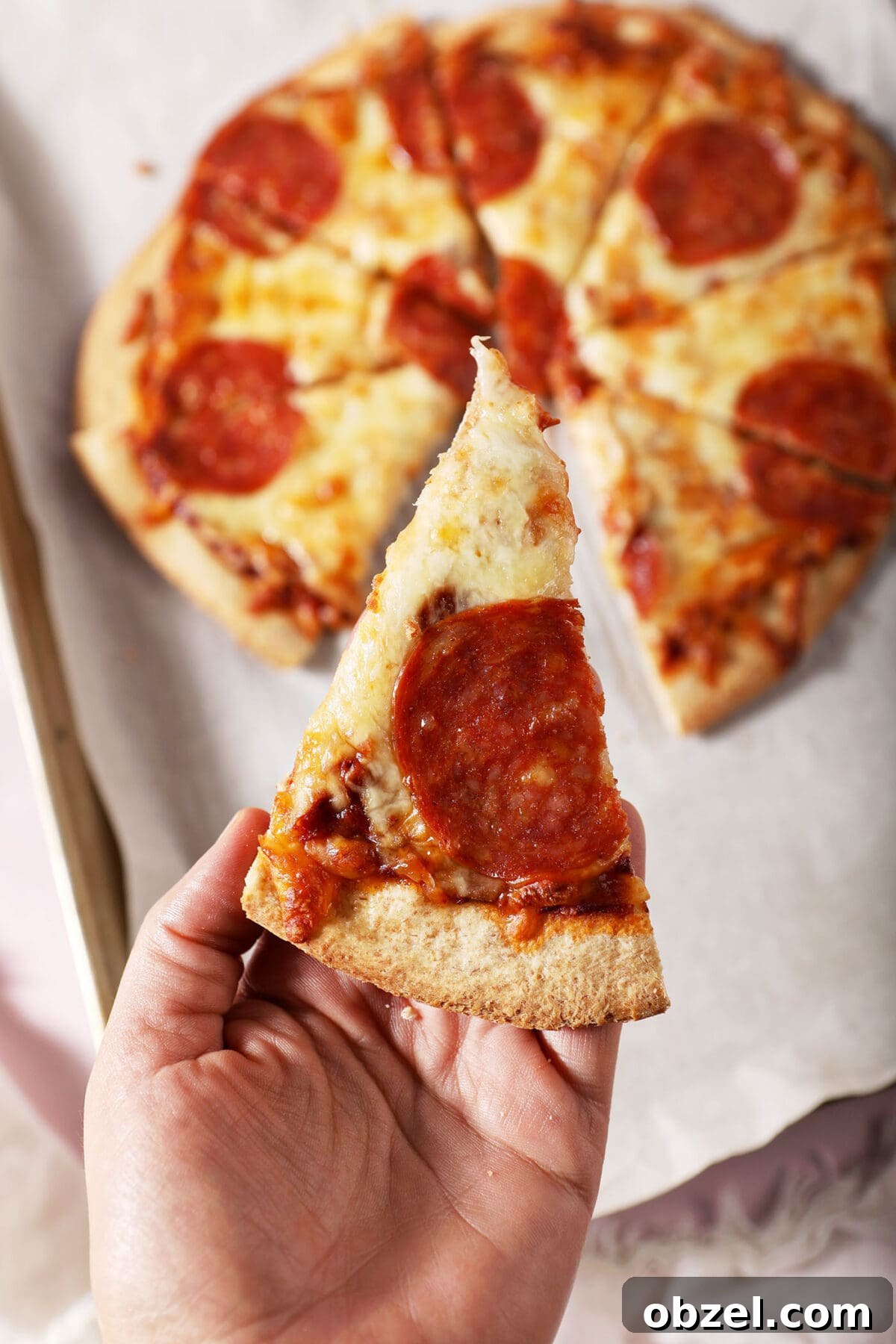 A hand holds a slice of homemade pepperoni pizza
