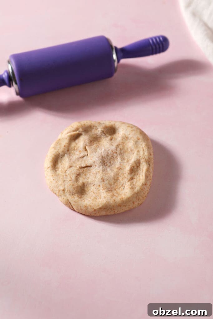 A round of pizza dough pressed down on a pink surface next to a purple rolling pin