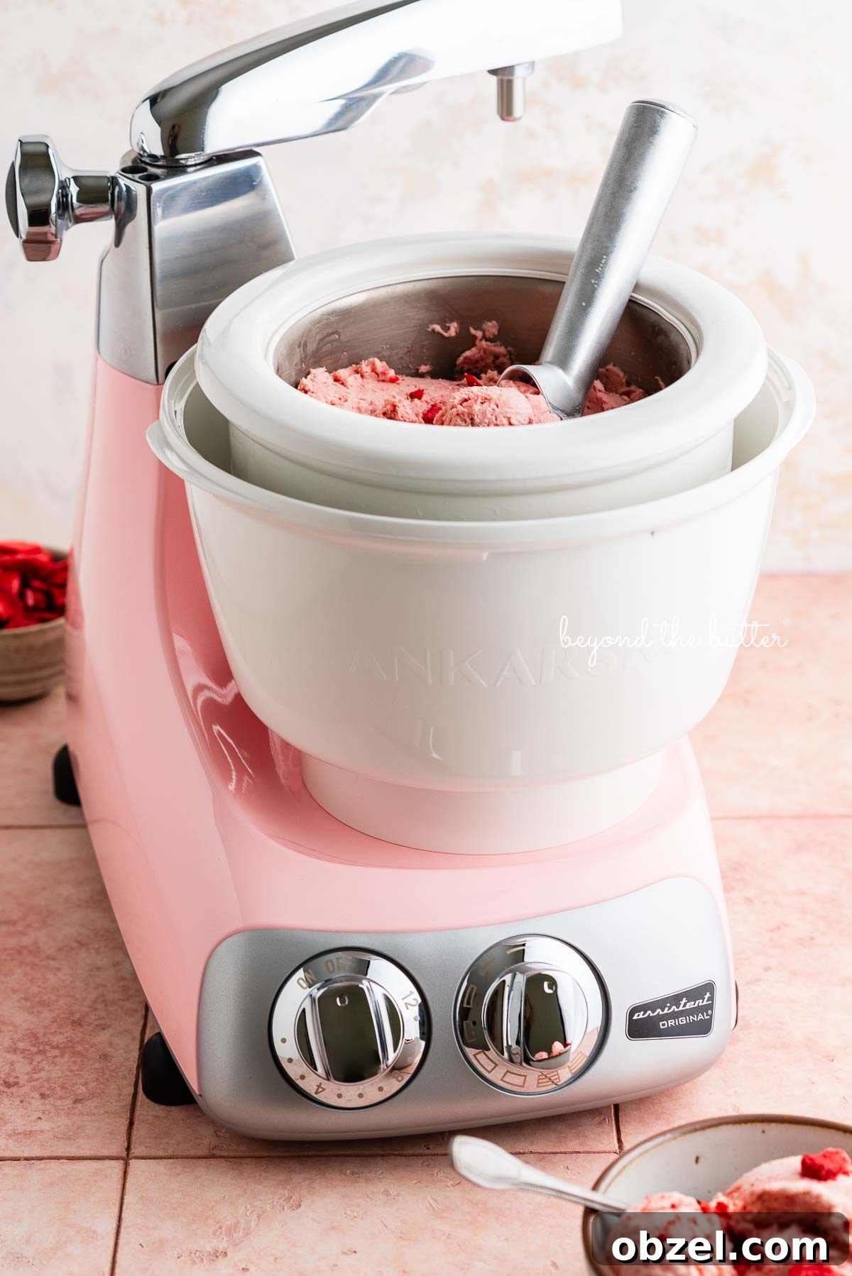 Cloudy pink Ankarsrum Assistent Original Mixer and ice cream maker attachment containing homemade strawberry ice cream with a bowl of ice cream and freeze-dried strawberries around it on a pink title background.