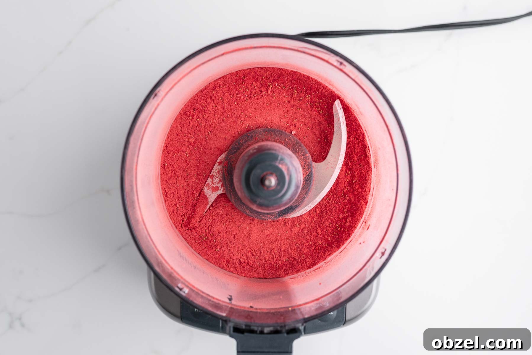 Processed freeze-dried strawberries in a medium-sized food processor on a white marbled background.