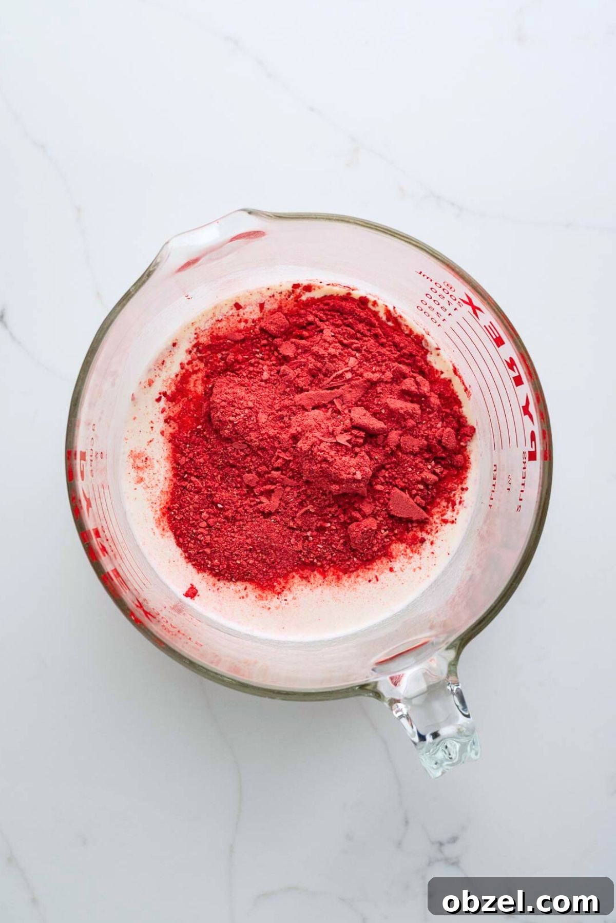Strawberry ice cream ingredients added to a large glass mixing bowl on a white marbled background.