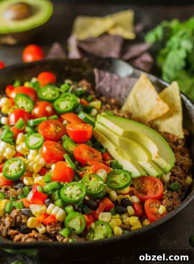 Close-up of a colorful skillet filled with healthy nachos topped with sliced avocado, cherry tomatoes, jalapeños, corn, black beans, and ground beef, served with tortilla chips.