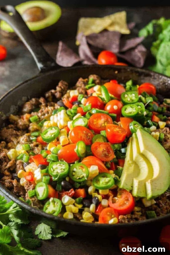 Side view of a cast iron pan with healthy nachos topped with fresh vegetables and avocado slices, surrounded by ingredients like cherry tomatoes, cilantro, and an avocado half.