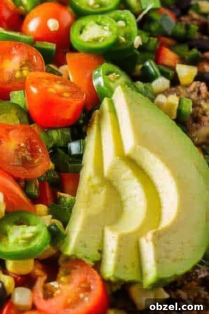 Extreme close-up of fresh avocado slices surrounded by chopped cherry tomatoes, jalapeños, green onions, and corn atop loaded healthy nachos.