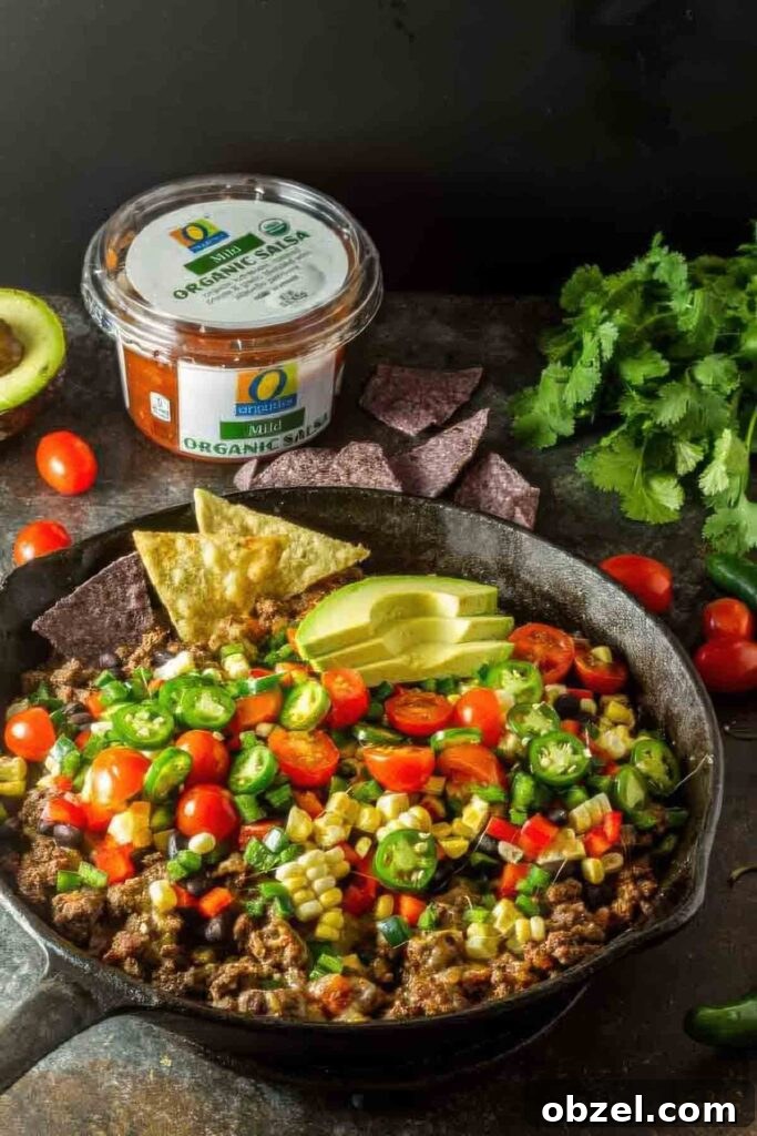 Overhead shot of a cast iron skillet filled with healthy nachos made with seasoned ground beef, fresh vegetables, and avocado, with blue corn and yellow tortilla chips on the side.