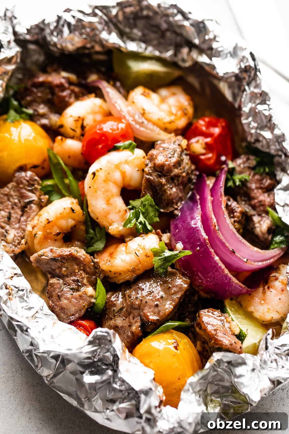 close up shot of a foil pack filled with steak bites, shrimp, red onions, and colored grape tomatoes.