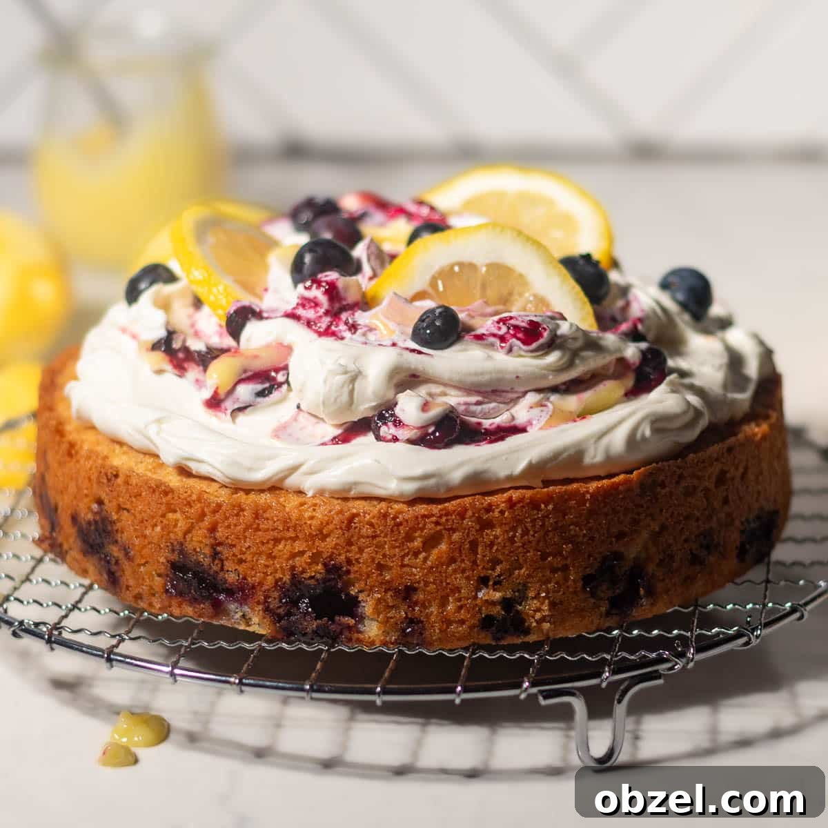 Lemon Blueberry Cake with Mascarpone Frosting 24 Full lemon blueberry mascarpone cake on a metal rack, topped with mascarpone cream, blueberries, and lemon slices.