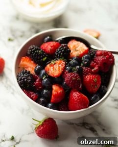 Berry Fruit Salad berries in small white bowl with silver spoon surrounded by strawberries on marble background