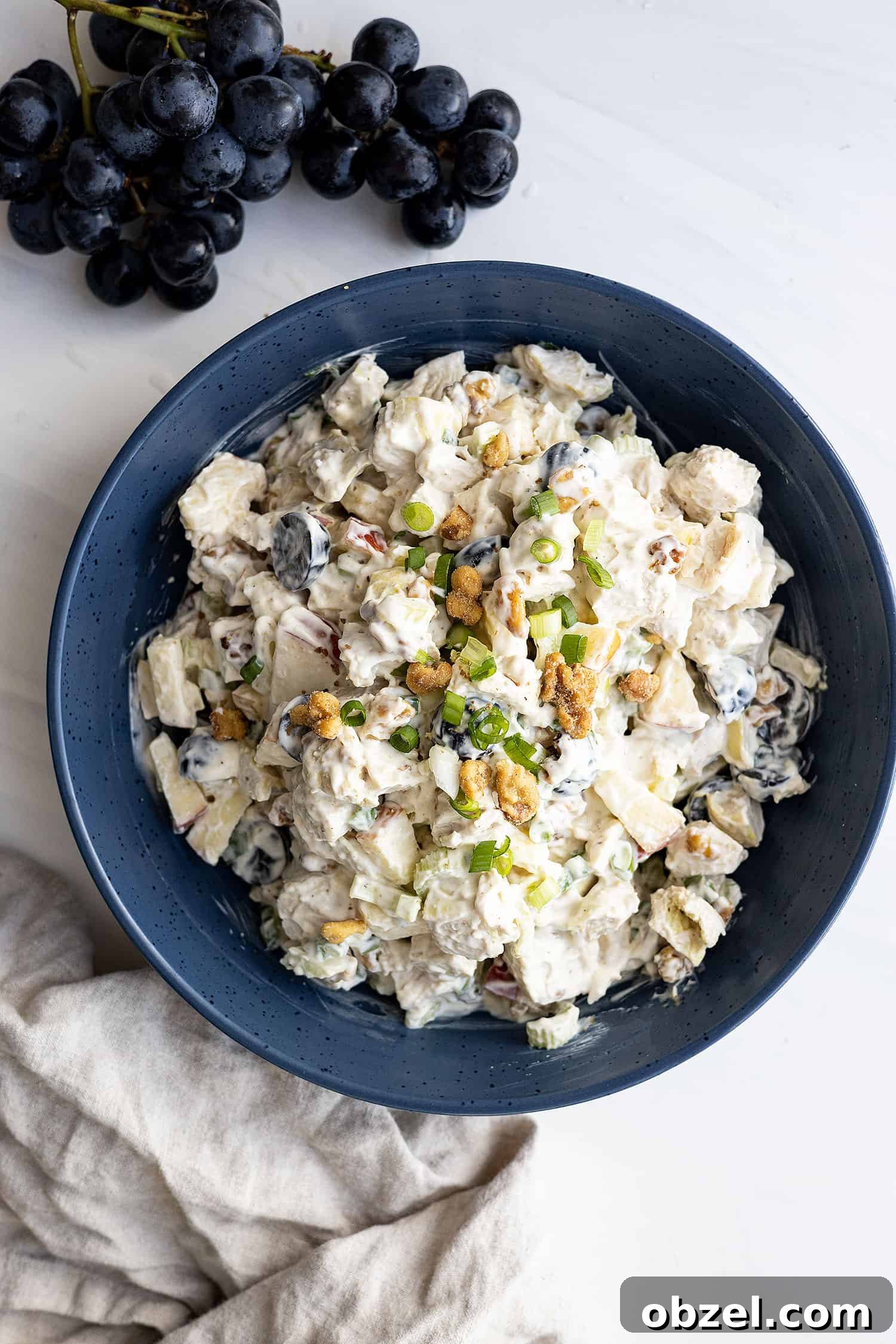 Chicken Waldorf Salad Recipe with Apples and Grapes 6 Overhead view of chicken waldorf salad in a large dark blue bowl garnished with chopped walnuts and green onion. Purple grapes in the corner and a light colored napkin beside the bowl.