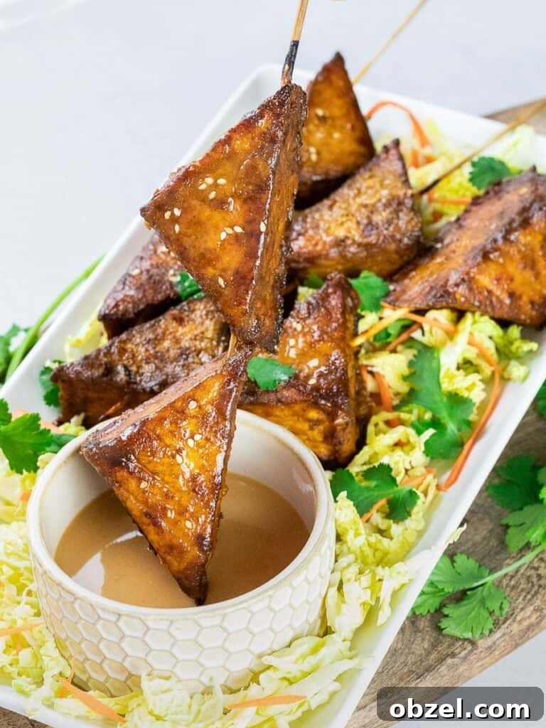 baked thai tofu satay skewers dipped in peanut satay sauce on a white plate with wooden board