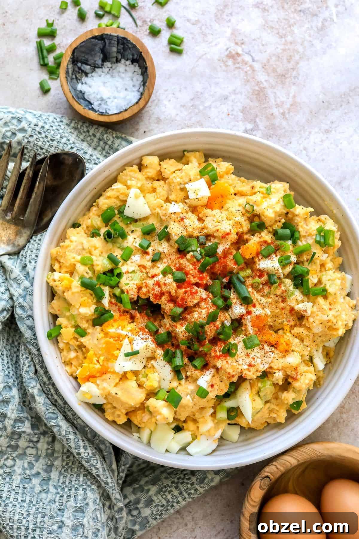 Deviled Egg Potato Salad Recipe 2 White ceramic bowl with deviled egg potato salad. Green onions on top and to the side with a spoon.