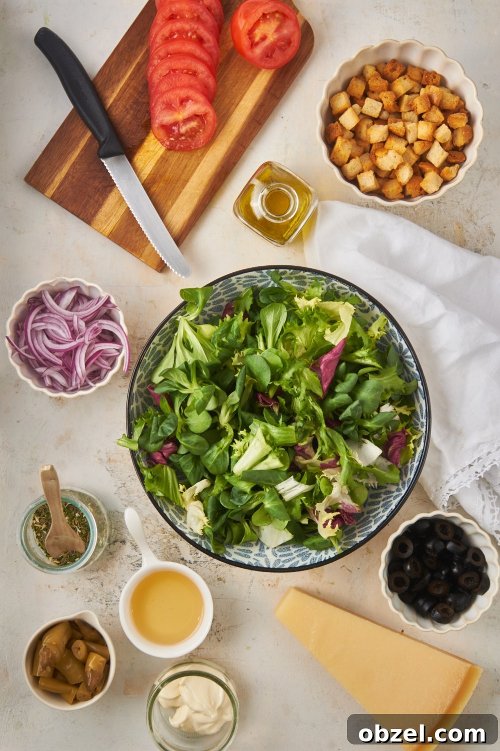Olive Garden Salad Recipe with Homemade Dressing 5 Overhead view of an assortment of ingredients including bowls of lettuce mix, croutons, sliced red onions, black olives, peppers, mayonnaise, a piece of parmesan cheese, sliced tomatoes on a cutting board, and a jar of oil.