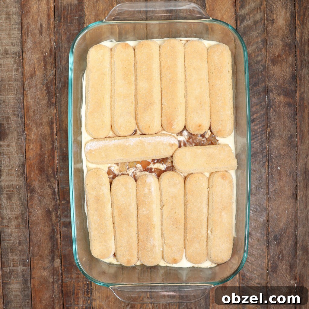 Caramel Apple Tiramisu Recipe: Autumn Dessert Twist 5 A glass baking dish filled with layers of ladyfinger cookies and creamy filling, arranged on a rustic wooden surface, showcases a delightful caramel apple tiramisu dessert in the making.