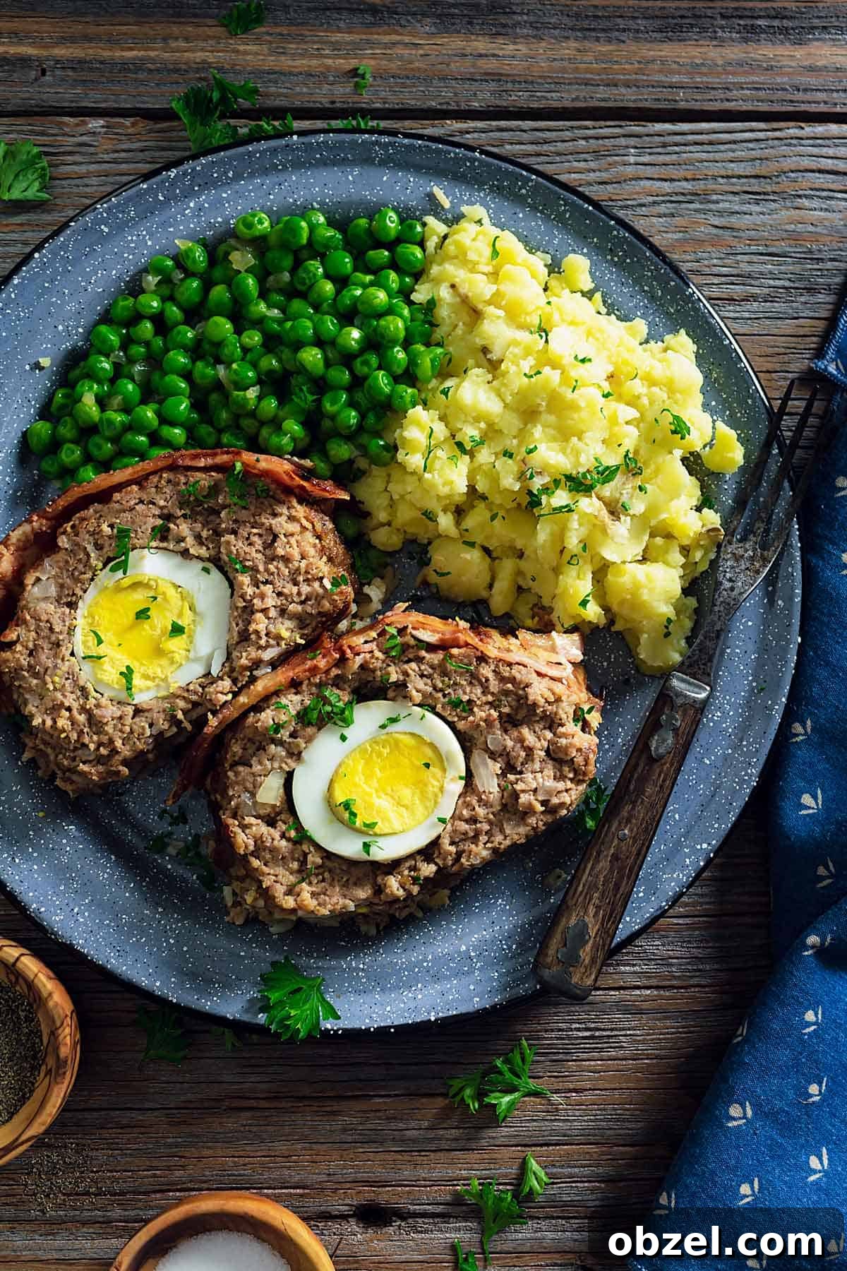 An overhead photo of two meatloaf slices showing the boiled eggs inside on a plate with mashed potatoes and green peas.