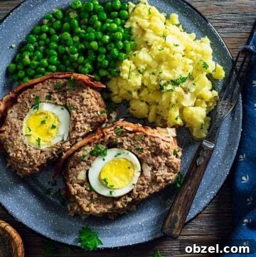 An overhead photo of two meatloaf slices showing the boiled eggs inside on a plate with mashed potatoes and green peas.