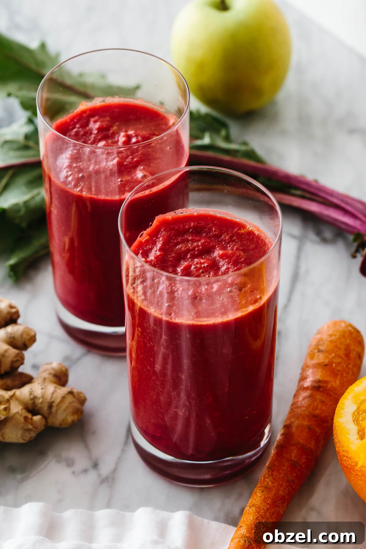 Two tall glasses of smoothie, surrounded by carrots, beets, apple and ginger.