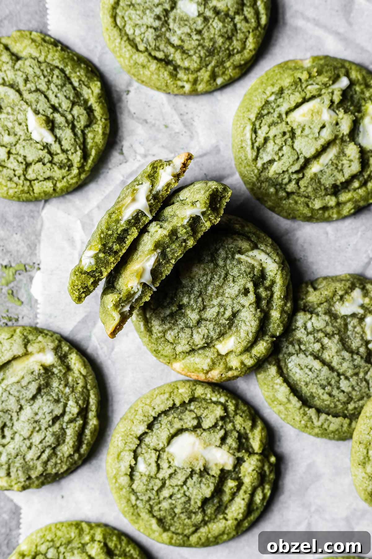 Matcha White Chocolate Cookies Recipe 10 A matcha white chocolate cookie is broken in half, revealing the gooey melted chocolate inside. There are many other matcha cookies surrounding it.