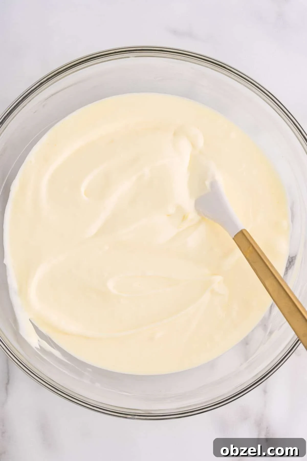 Creamy Loaf Pan Cheesecake Recipe 11 Overhead view of smooth, creamy cheesecake batter in a glass mixing bowl with a gold-handled spatula, ready to be poured into the pan