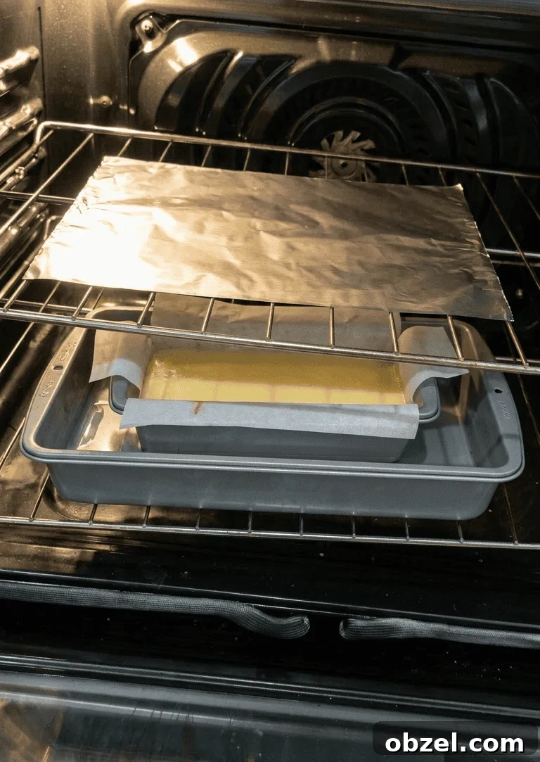 Creamy Loaf Pan Cheesecake Recipe 13 A loaf pan of cheesecake batter in a water bath inside a preheated oven, with a loose sheet of aluminum foil tented on the rack above to prevent overbrowning