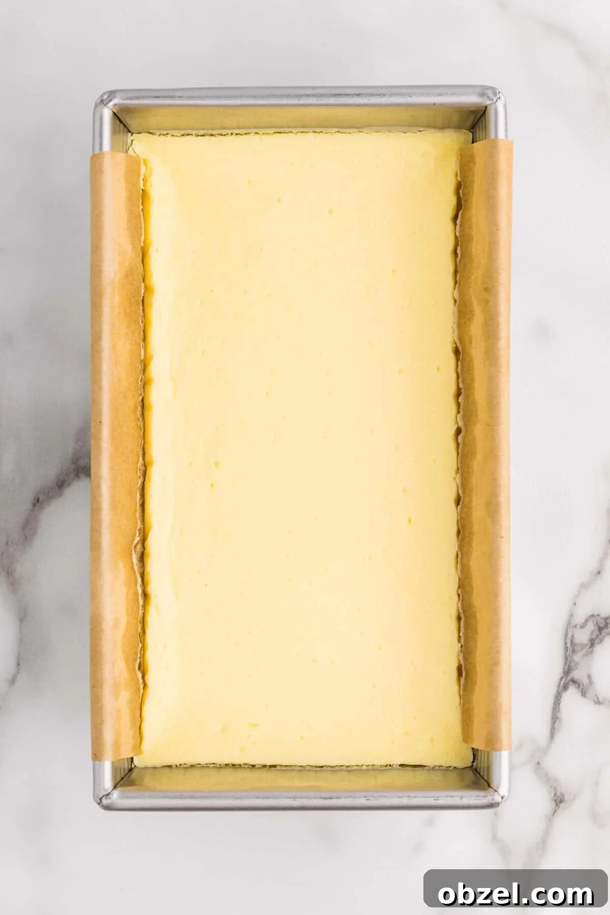 Creamy Loaf Pan Cheesecake Recipe 14 Overhead view of a fully baked cheesecake in a parchment-lined loaf pan, showing a smooth, pale yellow surface after baking