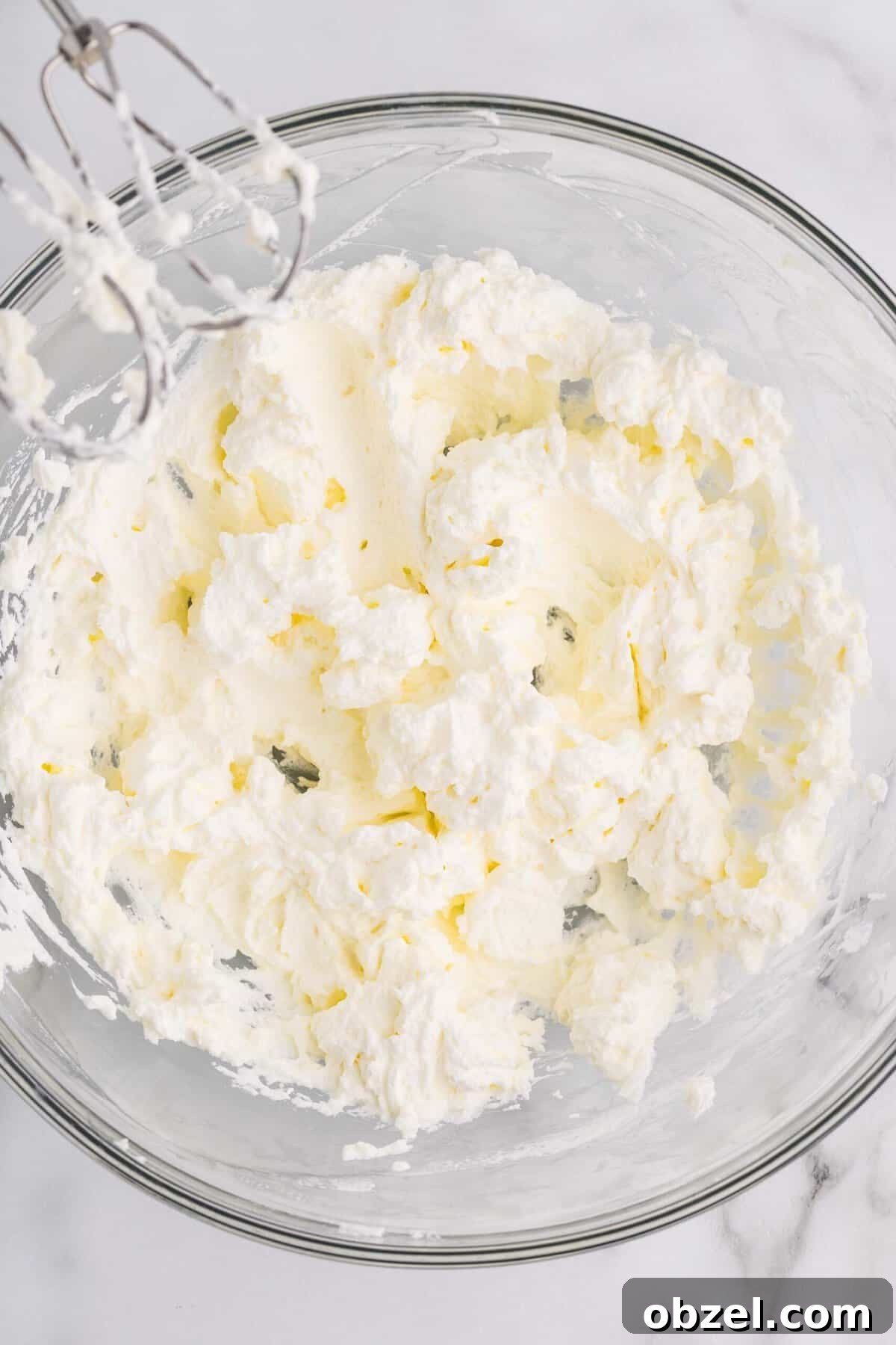 Creamy Loaf Pan Cheesecake Recipe 16 Overhead view of cream cheese being beaten with a hand mixer in a glass bowl, showing a fluffy and whipped texture at the start of making cheesecake filling