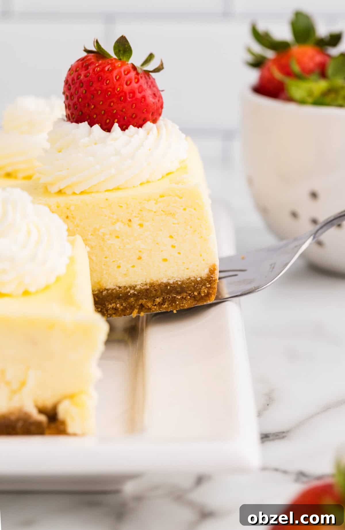 Creamy Loaf Pan Cheesecake Recipe 17 Close-up side view of a triangular loaf pan cheesecake slice topped with whipped cream and a fresh strawberry being lifted with a cake server, showing the smooth creamy filling and graham cracker crust