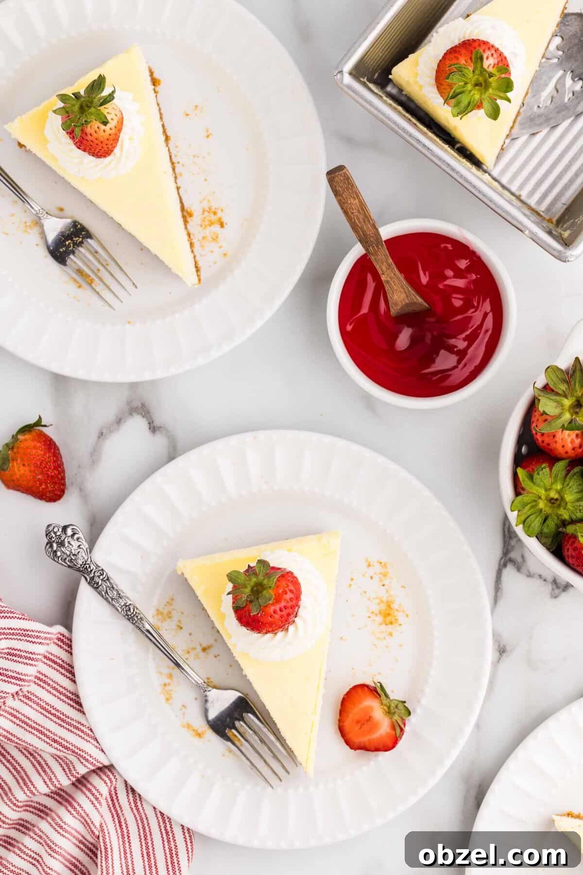 Creamy Loaf Pan Cheesecake Recipe 19 Overhead view of two white plates each with a triangular cheesecake slice topped with whipped cream and a strawberry, beside a bowl of strawberry sauce, fresh strawberries, and the remaining cheesecake in the loaf pan