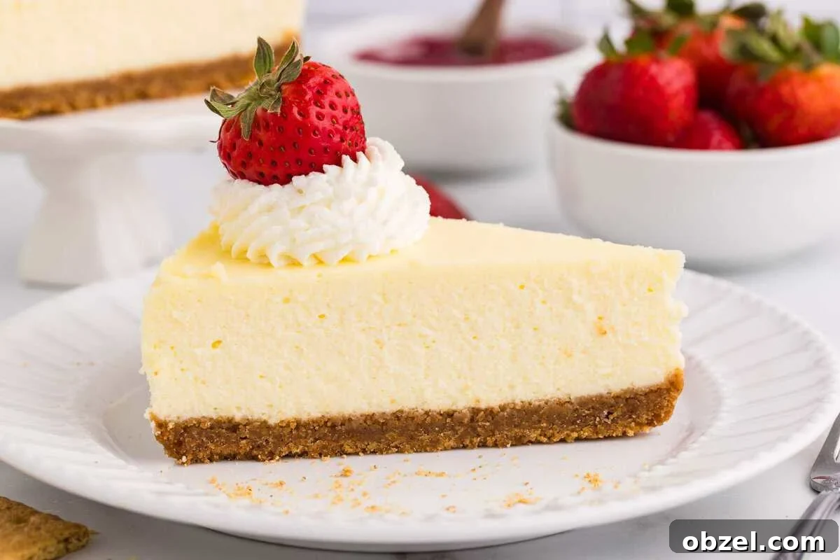Creamy Loaf Pan Cheesecake Recipe 5 Close-up of a creamy cheesecake slice with graham cracker crust, topped with whipped cream and a strawberry on a white plate, with the full cheesecake on a cake stand and fresh strawberries in a bowl in the background
