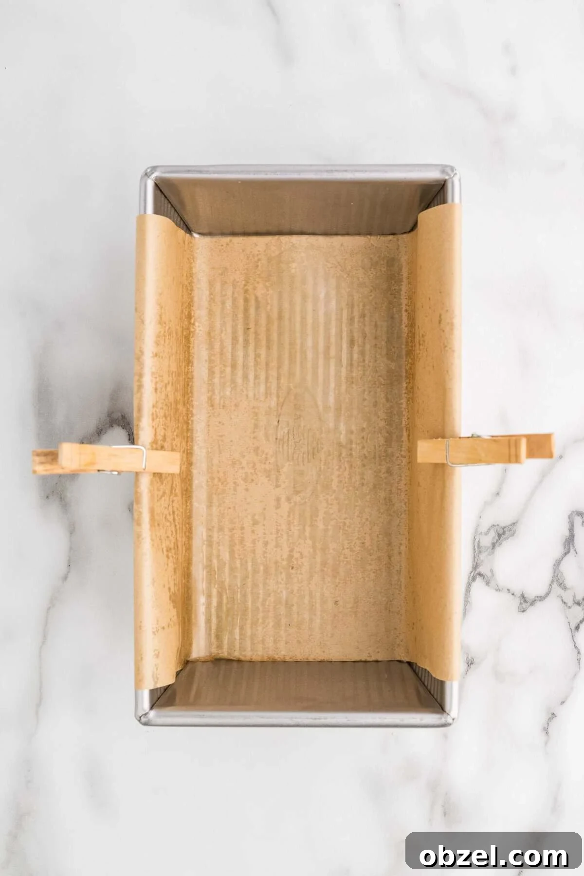 Creamy Loaf Pan Cheesecake Recipe 7 Overhead view of a loaf pan lined with parchment paper and secured with wooden clothespins, sitting on a marble surface