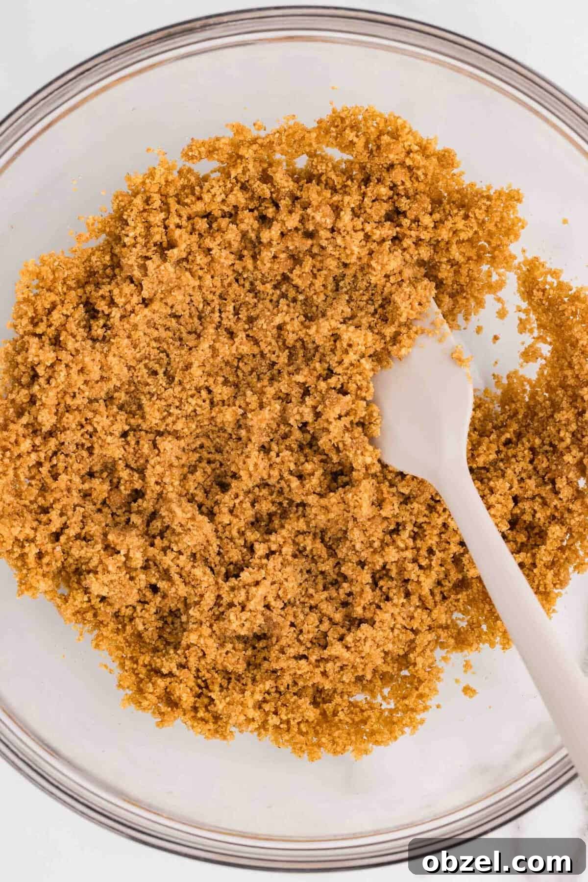 Creamy Loaf Pan Cheesecake Recipe 8 Overhead view of graham cracker crumbs mixed with melted butter in a glass mixing bowl, stirred with a white spoon to make cheesecake crust
