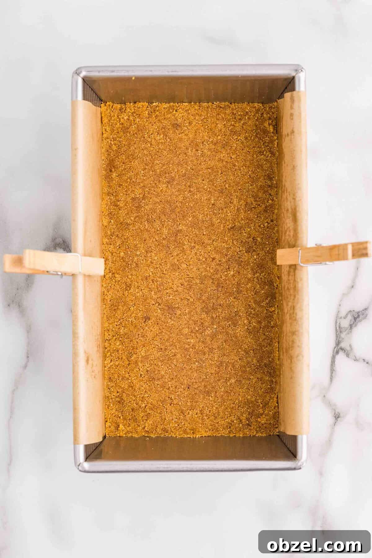 Creamy Loaf Pan Cheesecake Recipe 9 Overhead view of graham cracker crust pressed evenly into the bottom of a parchment-lined loaf pan secured with clothespins on a marble surface