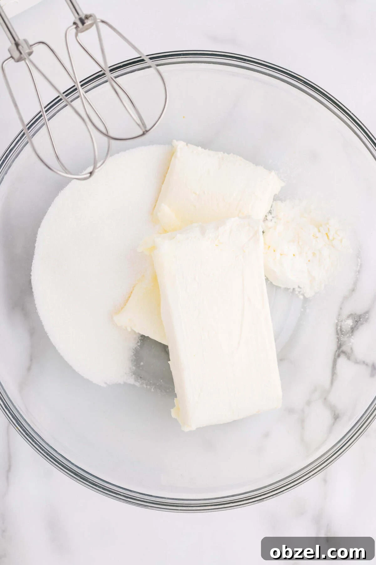 Creamy Loaf Pan Cheesecake Recipe 10 Overhead view of cream cheese blocks and granulated sugar in a glass mixing bowl with a hand mixer, ready to beat together for cheesecake filling