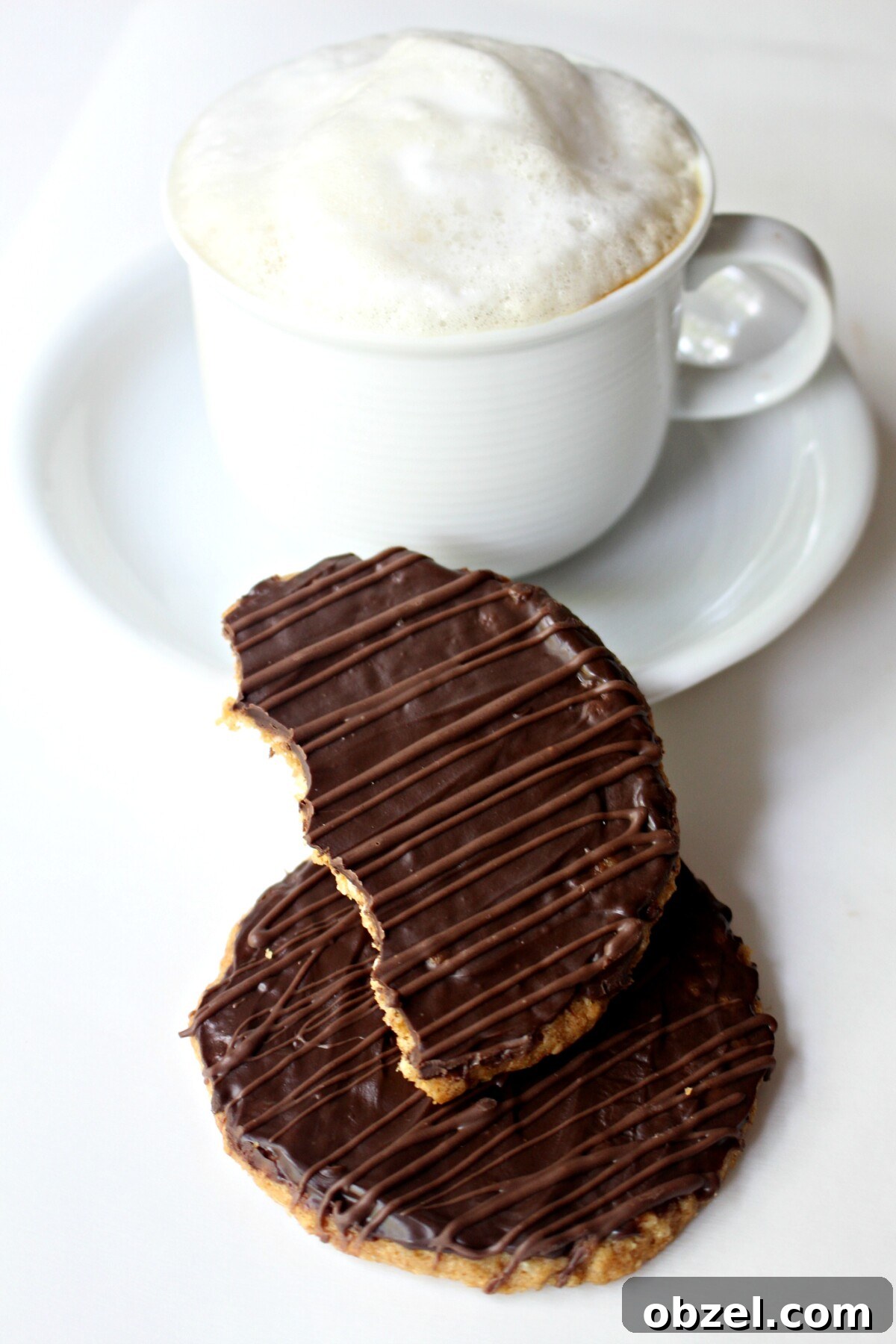 Chocolate Covered Hobnob Oat Cookies Recipe 7 Two cookies in front of a white mug of cappuccino topped with milk foam.