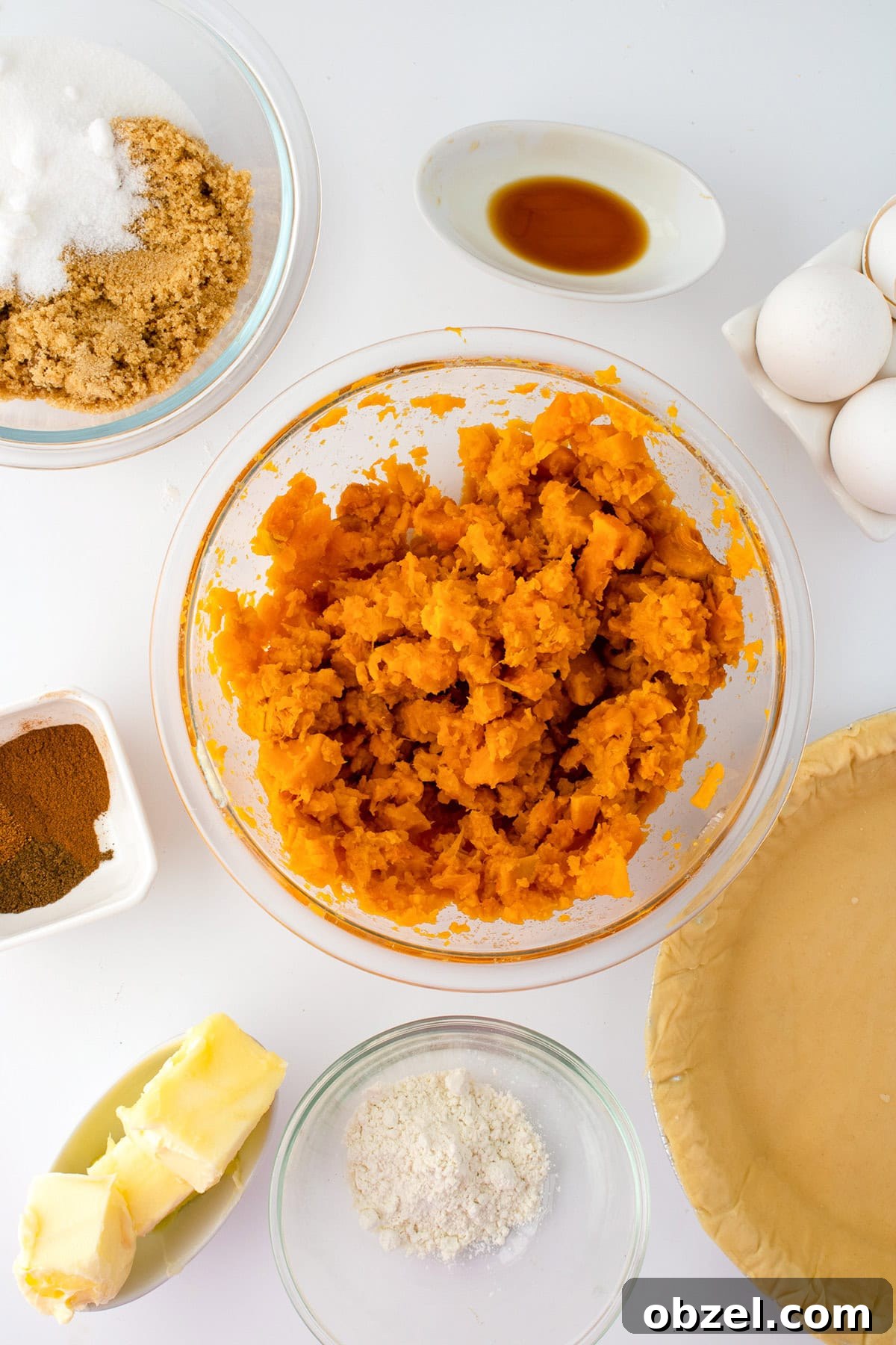 Overhead view of sweet potato pie ingredients, including mashed sweet potato, browned butter, sugar, eggs, pie crust, spices, flour, and vanilla extract on a white surface.